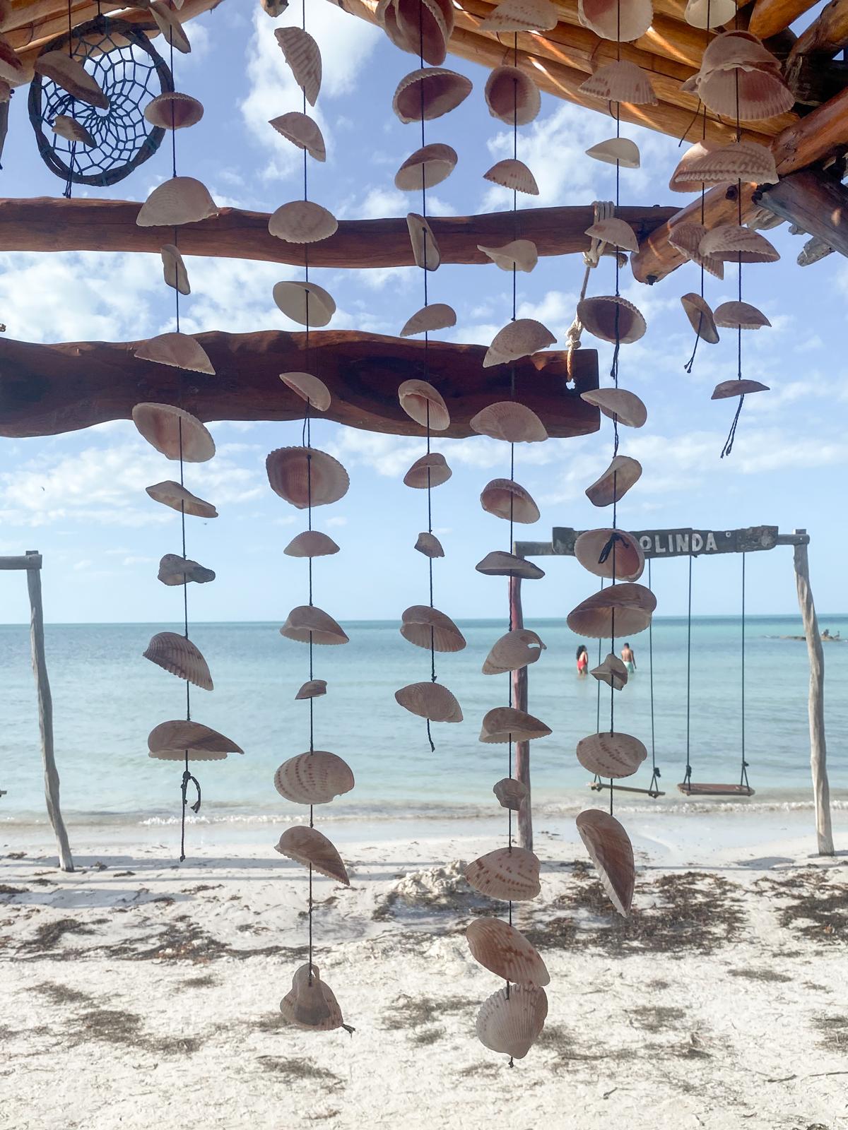 Hanging shell mobiles under a driftwood shelter above a white beach
